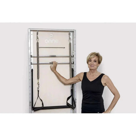 Femme posant à côté Pilates Wall Unit 5-en-1 montée au mur mettant en échelle humaine et accès aux barres métalliques