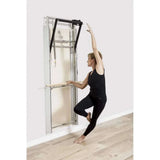Femme utilisant barre de ballet en bois clair sur Pilates Wall Unit 5-en-1 pour exercice d'équilibre à une jambe